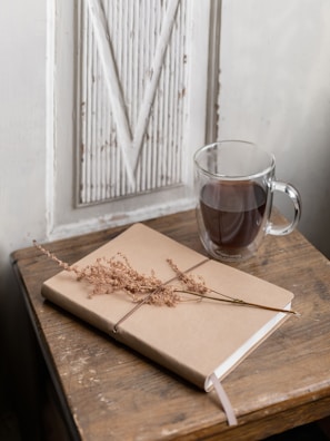 A minimalist setting featuring a closed journal with a tan cover and a dried flower on top, placed on a rustic wooden table. Next to the journal is a clear glass mug filled with dark coffee. The backdrop includes a white paneled section with a vintage look.