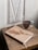 A minimalist setting featuring a closed journal with a tan cover and a dried flower on top, placed on a rustic wooden table. Next to the journal is a clear glass mug filled with dark coffee. The backdrop includes a white paneled section with a vintage look.