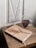 A minimalist setting featuring a closed journal with a tan cover and a dried flower on top, placed on a rustic wooden table. Next to the journal is a clear glass mug filled with dark coffee. The backdrop includes a white paneled section with a vintage look.