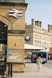 A historic railway station with a prominent brick structure featuring a large British Rail logo. A sign below provides parking directions, and a vintage clock hangs from the far wall. A modern blue and red bus marked with the number 26 is parked near the entrance. A person in summery clothing is walking along the pavement.