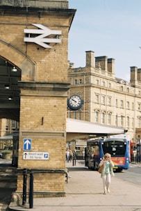 A historic railway station with a prominent brick structure featuring a large British Rail logo. A sign below provides parking directions, and a vintage clock hangs from the far wall. A modern blue and red bus marked with the number 26 is parked near the entrance. A person in summery clothing is walking along the pavement.