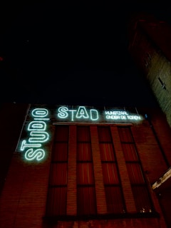A night view of a building facade illuminated by neon signs. The signs display the words 'STAD' and 'STUDIO' in large, glowing letters, with additional smaller text 'KUNSTHAL ONDER DE TOREN' also lit up. The building features tall, vertical windows with red lighting seen through them.