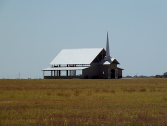 A solitary church building with a tall steeple stands in the middle of an expansive grassy field under a clear blue sky. The architecture displays a simple, traditional design with a white roof and open sections under construction.