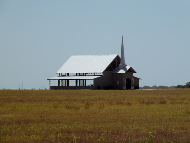 A solitary church building with a tall steeple stands in the middle of an expansive grassy field under a clear blue sky. The architecture displays a simple, traditional design with a white roof and open sections under construction.