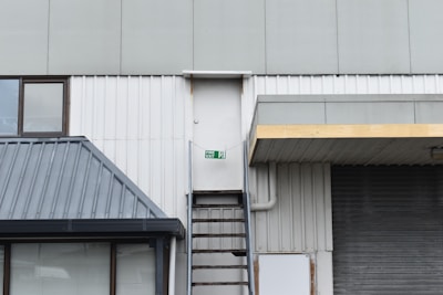 A white door is situated high above the ground on the side of an industrial building, with a metal ladder leading up to it. There is a green fire exit sign above the door. The building exterior includes various metal and corrugated surfaces with a muted color palette. A tinted window is visible on the left side of the image, framed by dark metal.