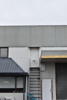A building exterior features a metal fire escape ladder leading up to a white door with a fire exit sign. The structure includes corrugated metal and a slanted roof over a window. Adjacent to the ladder, there is a closed roller shutter and a standard door.