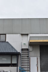 A building exterior features a metal fire escape ladder leading up to a white door with a fire exit sign. The structure includes corrugated metal and a slanted roof over a window. Adjacent to the ladder, there is a closed roller shutter and a standard door.