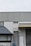 A building exterior features a metal fire escape ladder leading up to a white door with a fire exit sign. The structure includes corrugated metal and a slanted roof over a window. Adjacent to the ladder, there is a closed roller shutter and a standard door.