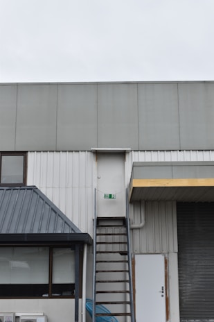 A building exterior features a metal fire escape ladder leading up to a white door with a fire exit sign. The structure includes corrugated metal and a slanted roof over a window. Adjacent to the ladder, there is a closed roller shutter and a standard door.