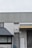 A building exterior features a metal fire escape ladder leading up to a white door with a fire exit sign. The structure includes corrugated metal and a slanted roof over a window. Adjacent to the ladder, there is a closed roller shutter and a standard door.