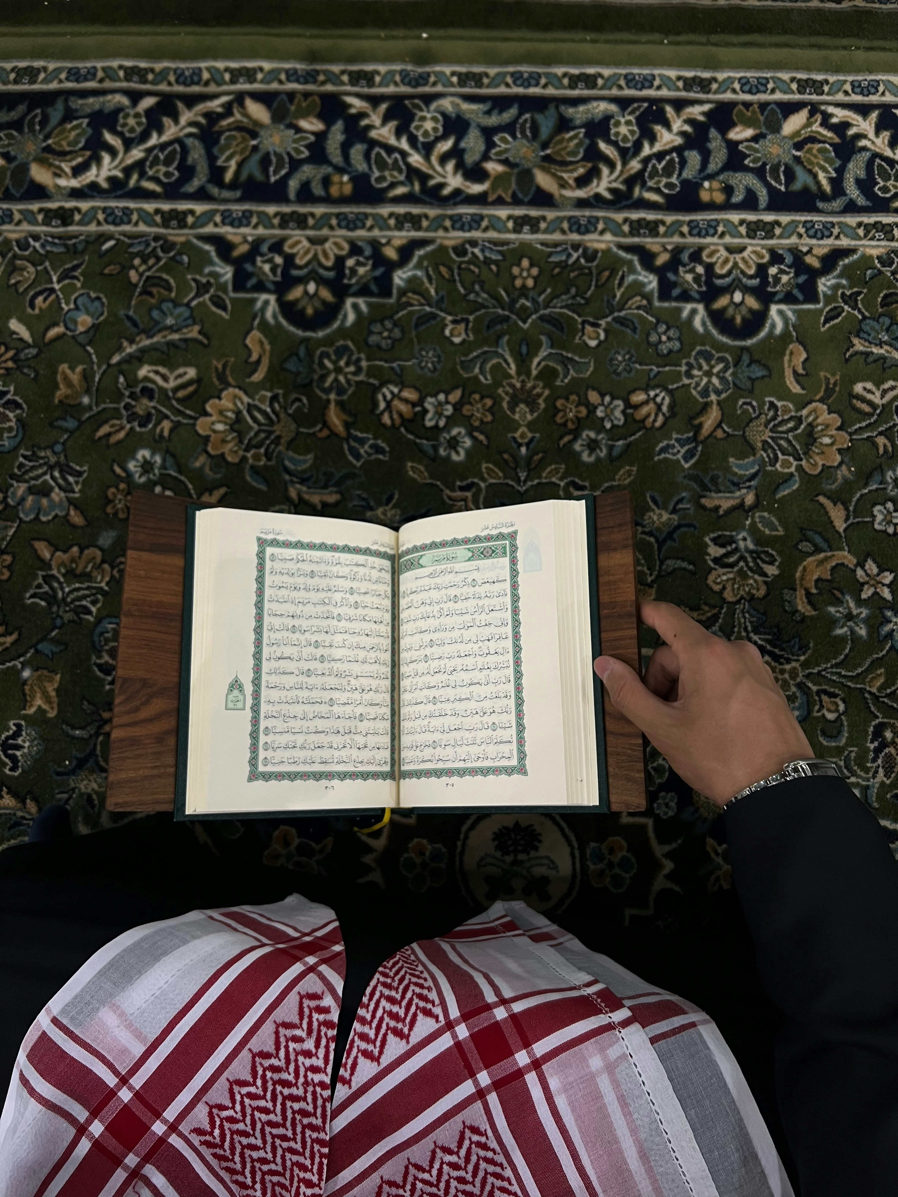An open book with Arabic script is held by a person wearing traditional attire. The book rests on what appears to be a lectern or stand. The person is dressed in a black garment with a red and white patterned keffiyeh. The background features a detailed, floral-patterned carpet in shades of green, blue, and beige.