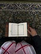 An open book with Arabic script is held by a person wearing traditional attire. The book rests on what appears to be a lectern or stand. The person is dressed in a black garment with a red and white patterned keffiyeh. The background features a detailed, floral-patterned carpet in shades of green, blue, and beige.