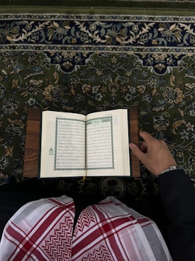 An open book with Arabic script is held by a person wearing traditional attire. The book rests on what appears to be a lectern or stand. The person is dressed in a black garment with a red and white patterned keffiyeh. The background features a detailed, floral-patterned carpet in shades of green, blue, and beige.