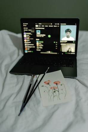 A laptop displays a music streaming interface, resting on a white bed. In front of the laptop is a card featuring an art piece of red flowers with three paintbrushes beside it.