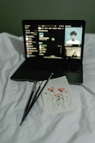 A laptop displays a music streaming interface, resting on a white bed. In front of the laptop is a card featuring an art piece of red flowers with three paintbrushes beside it.