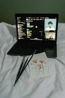 A laptop displays a music streaming interface, resting on a white bed. In front of the laptop is a card featuring an art piece of red flowers with three paintbrushes beside it.