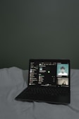 A laptop is open on a bed with white sheets, displaying a music streaming application. The screen shows a playlist and track details, with album cover art visible on the right side.