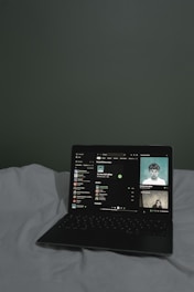 A laptop is open on a bed with white sheets, displaying a music streaming application. The screen shows a playlist and track details, with album cover art visible on the right side.