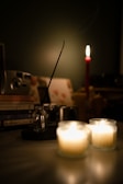 A dimly lit scene featuring two small candles in glass holders, casting a warm and soft light. An incense stick burns in a small glass bottle, with a faint trail of smoke visible. In the background, there's a red candle standing upright, while various objects like books, a camera, and a blurred frame create a cozy and serene atmosphere.