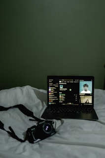 A dimly lit scene featuring a laptop placed on a white bedspread. The laptop screen displays a music streaming interface with album art and track information. Next to the laptop, there is a camera with a strap lying on the bed.