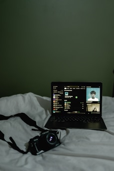 A dimly lit scene featuring a laptop placed on a white bedspread. The laptop screen displays a music streaming interface with album art and track information. Next to the laptop, there is a camera with a strap lying on the bed.