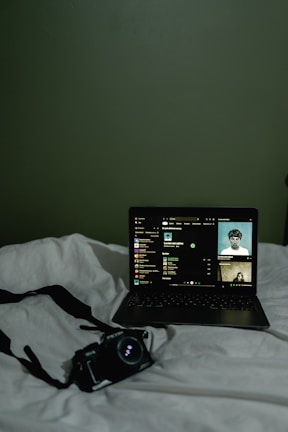 A dimly lit scene featuring a laptop placed on a white bedspread. The laptop screen displays a music streaming interface with album art and track information. Next to the laptop, there is a camera with a strap lying on the bed.