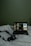 A dimly lit scene featuring a laptop placed on a white bedspread. The laptop screen displays a music streaming interface with album art and track information. Next to the laptop, there is a camera with a strap lying on the bed.