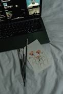 A laptop on a bed displays a music streaming platform with album art and song details. Next to it are a few paintbrushes and a card featuring a watercolor painting of pink flowers and green stems.