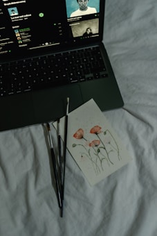 A laptop on a bed displays a music streaming platform with album art and song details. Next to it are a few paintbrushes and a card featuring a watercolor painting of pink flowers and green stems.