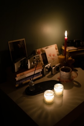 A dimly lit tabletop displays two lit candles casting soft glows. A red taper candle burns in the background, surrounded by stacked books and several vintage cameras. A steaming mug sits next to an incense holder, with photographs leaning against the wall.