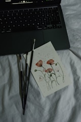 A close-up of a painting with delicate red flowers on a piece of paper lying on a soft, white surface. Nearby are three paintbrushes with dark handles, and the corner of an open laptop keyboard is visible, displaying a music player interface on the screen.