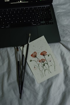 A close-up of a painting with delicate red flowers on a piece of paper lying on a soft, white surface. Nearby are three paintbrushes with dark handles, and the corner of an open laptop keyboard is visible, displaying a music player interface on the screen.