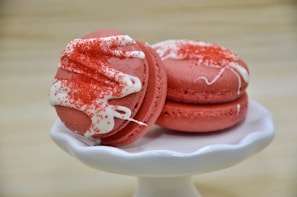 Two pink macarons, embellished with white icing and red sprinkles, are neatly arranged on a small white pedestal stand. The background is softly blurred, drawing attention to the vibrant desserts.