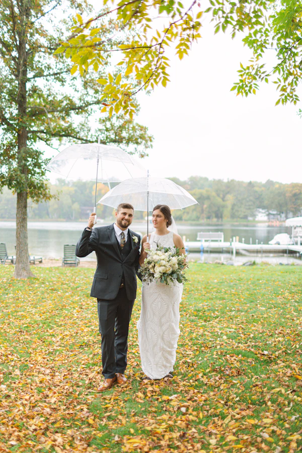 A bride and groom walking under a clear umbrella on a leaf-covered lawn by a lake
