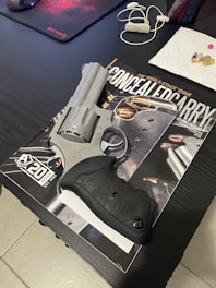 A silver revolver sits on top of a magazine titled 'Concealed Carry'. Nearby are a mousepad, earbuds, and several pills on a paper towel, all placed on a dark table.