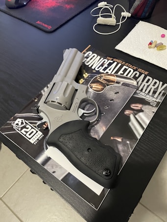 A silver revolver sits on top of a magazine titled 'Concealed Carry'. Nearby are a mousepad, earbuds, and several pills on a paper towel, all placed on a dark table.