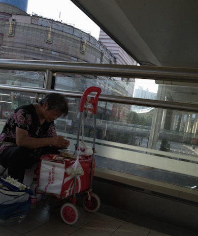 An elderly person is seated on a two-wheeled trolley in an urban environment next to a high wall of windows. They are engaged in an activity with their hands, possibly preparing or eating food. The scene includes a red trolley with various items attached, such as a bag. A large building is visible outside the window.