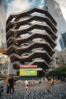 A large, spiral architectural structure with multiple levels stands prominently in an urban setting. In front of the structure, a large screen displays a sports broadcast, and several people are gathered around, some sitting and others standing. The scene is set outdoors, with towering modern buildings surrounding the area. The ground is paved with a geometric pattern of gray and white tiles.