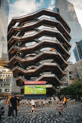 A large, spiral architectural structure with multiple levels stands prominently in an urban setting. In front of the structure, a large screen displays a sports broadcast, and several people are gathered around, some sitting and others standing. The scene is set outdoors, with towering modern buildings surrounding the area. The ground is paved with a geometric pattern of gray and white tiles.