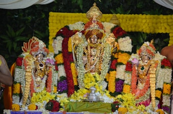 A richly adorned altar featuring three ornate statues, draped with vibrant flower garlands in shades of yellow, orange, red, purple, and white. The central statue is larger and heavily decorated with gold jewelry and intricate designs. The surrounding green foliage provides a lush background, enhancing the colorful and festive appearance.