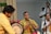 A group of musicians is performing, featuring a man in a yellow shirt playing a drum and another in a yellow traditional outfit playing a wind instrument. Additional musicians and individuals can be seen in the background, dressed in white with traditional decorations.