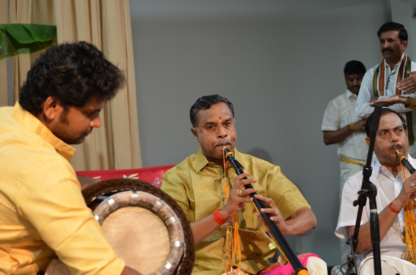 A group of musicians is performing, featuring a man in a yellow shirt playing a drum and another in a yellow traditional outfit playing a wind instrument. Additional musicians and individuals can be seen in the background, dressed in white with traditional decorations.