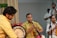 A group of musicians is performing, featuring a man in a yellow shirt playing a drum and another in a yellow traditional outfit playing a wind instrument. Additional musicians and individuals can be seen in the background, dressed in white with traditional decorations.