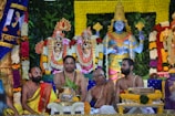 A colorful and elaborate Hindu religious ceremony featuring statues of deities adorned with floral garlands in varying colors. Four men are seated in front of the statues, wearing traditional Indian clothing and markings on their foreheads, indicating a devotional context. The background is filled with lush greenery and ornamental decorations.