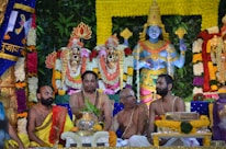 A colorful and elaborate Hindu religious ceremony featuring statues of deities adorned with floral garlands in varying colors. Four men are seated in front of the statues, wearing traditional Indian clothing and markings on their foreheads, indicating a devotional context. The background is filled with lush greenery and ornamental decorations.