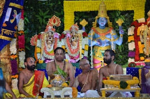 A colorful and elaborate Hindu religious ceremony featuring statues of deities adorned with floral garlands in varying colors. Four men are seated in front of the statues, wearing traditional Indian clothing and markings on their foreheads, indicating a devotional context. The background is filled with lush greenery and ornamental decorations.