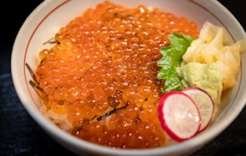 A bowl of rice topped with vibrant orange salmon roe, accompanied by a garnish of green wasabi, pale yellow ginger, fresh green shiso leaf, and a slice of red and white radish.