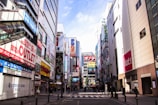 A bustling city street lined with tall buildings adorned with numerous colorful billboards and advertisements. People can be seen walking along the sidewalks, and the scene conveys an atmosphere of commercial vibrancy.