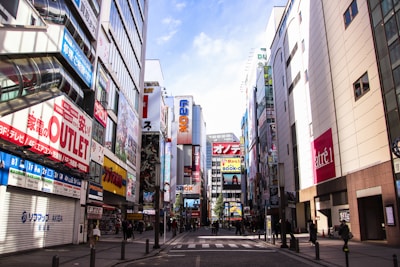 A bustling city street lined with tall buildings adorned with numerous colorful billboards and advertisements. People can be seen walking along the sidewalks, and the scene conveys an atmosphere of commercial vibrancy.
