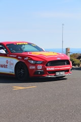 A red and yellow racing car, adorned with various sponsor logos, is parked on an asphalt road. It features a sleek design with sporty accents and a visible license plate. The background shows a clear blue sky and distant landscape.